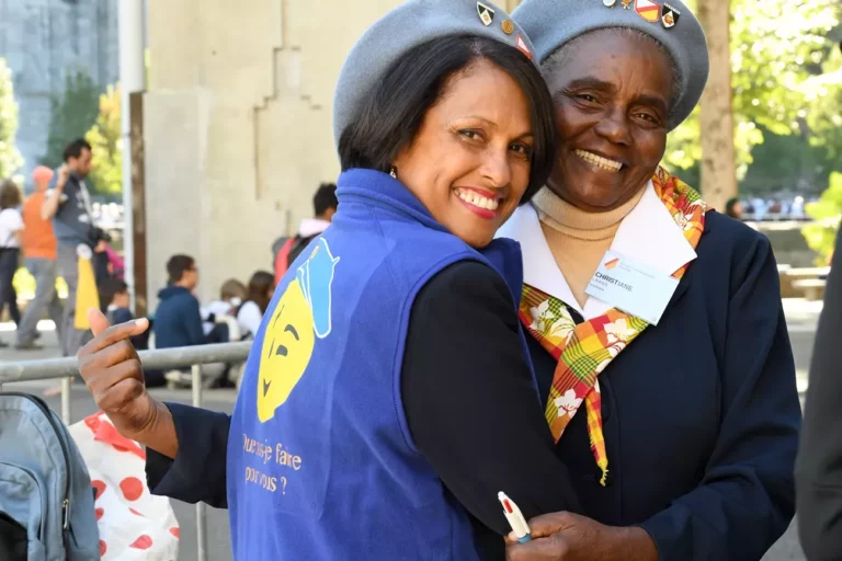 Deux femmes souriantes en bérets gris s'étreignant chaleureusement, l'une portant un gilet "Que puis-je faire pour vous ?"