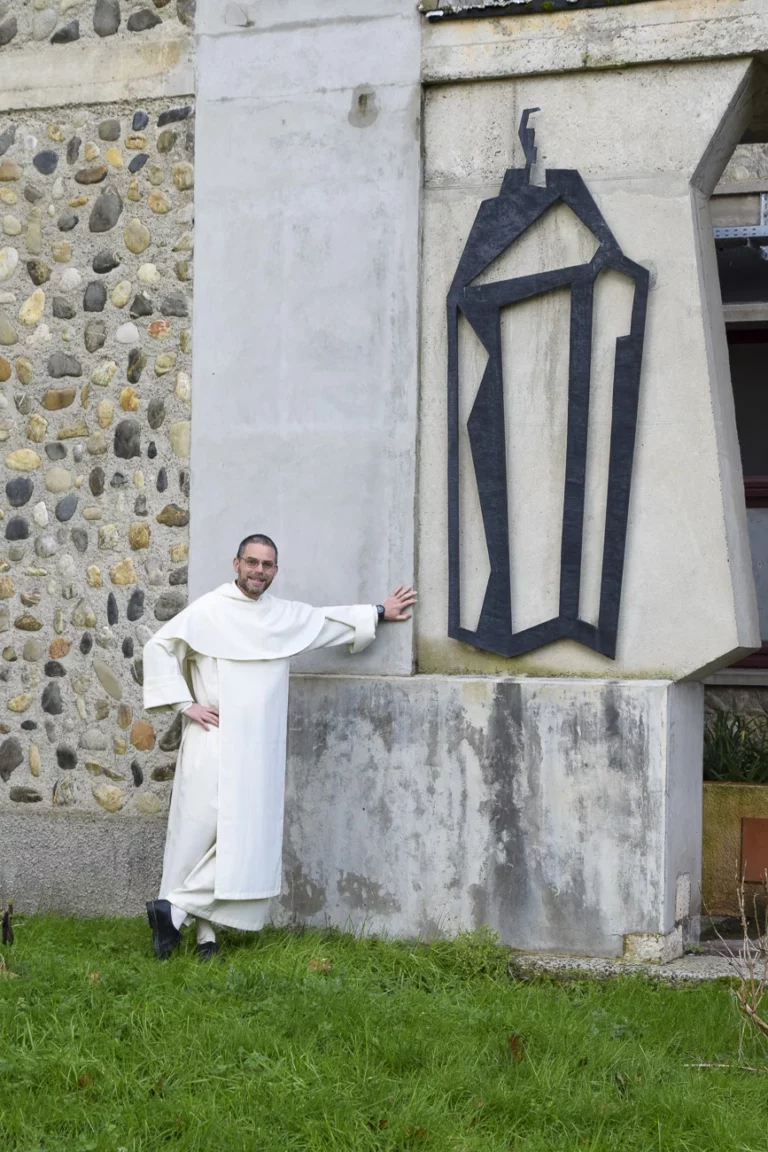 Moine souriant en habit blanc et lunettes, adossé à un mur d'abbaye avec une sculpture abstraite moderne, près d'un mur en galets.