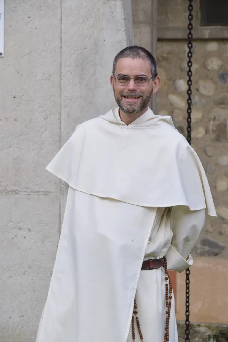 Portrait d'un moine souriant avec des lunettes et une barbe, vêtu d'un habit monastique blanc cassé et d'un chapelet en bois, devant un mur en pierre.