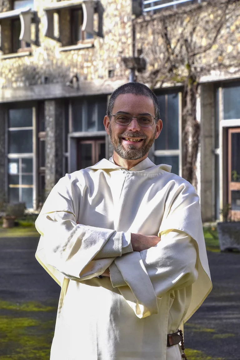 Portrait d'un moine barbu et souriant, portant des lunettes, un habit monastique clair et un chapelet, posant les bras croisés devant un monastère en pierre.
